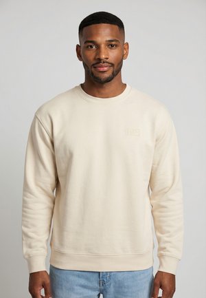 Uomo con capelli corti e barba che indossa una felpa color crema e jeans azzurri, rivolto in avanti su uno sfondo grigio uniforme.