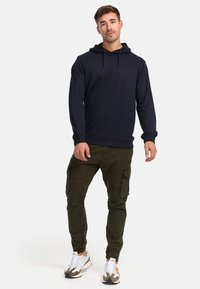 Marineblå hættetrøje med snøre og kængurulomme, parret med olivenfarvede cargobukser med sidelommer, og hvide sneakers med gule detaljer.