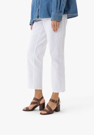 Person trägt weiße, gekürzte Hosen, braune Sandalen mit Blockabsatz und Schnallenriemen sowie ein blaues Jeanshemd und steht vor einem weißen Hintergrund.