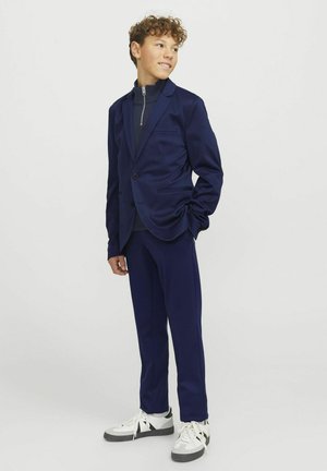 Costume marine comprenant un blazer à un bouton avec des revers en crans et un pantalon assorti. Associé à un turtleneck sombre et des baskets élégantes.