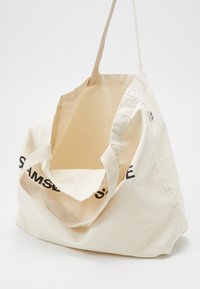 Tote bag en toile écrue avec de larges anses et un texte partiel noir imprimé sur le côté, présenté ouvert et vide sur un fond uni.