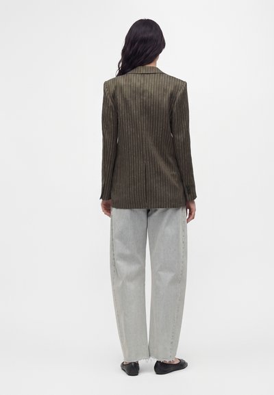 Femme de dos portant un blazer marron à fines rayures, un jean large gris clair et des chaussures plates noires sur fond blanc.