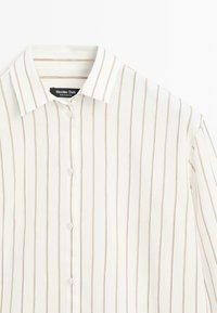 Witte blouse met dunne verticale beige strepen en een klassieke kraag, plat gelegd met het merklipje zichtbaar aan de binnenkant van de kraag.