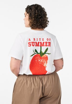 Kvinde med krøllet hår iført en hvid t-shirt med en bidt rød tomat og teksten "Et bid af sommer" på ryggen.
