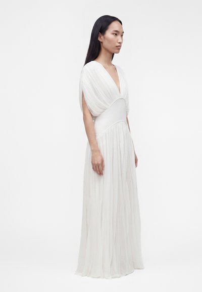 Γυναίκα με μακριά μαύρα μαλλιά φοράει μια αέρινη λευκή τουαλέτα με βαθύ ντεκολτέ σε σχήμα V και ρεβάνς, στέκεται μπροστά σε απλό λευκό φόντο.