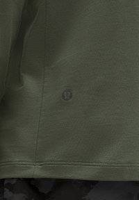 Camicia a maniche corte verde oliva realizzata in tessuto testurizzato, con un logo discreto sull'orlo inferiore e bordi cuciti in modo pulito.