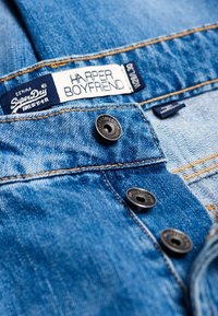 Modrá denimová látka s několika knoflíky, štítek s nápisem "SuperDry HARPER BOYFRIEND" a kontrastní prošití podél švů.