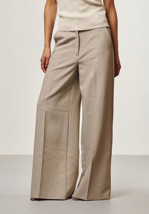 Vrouw die beige wijde broeken en een mouwloze beige top draagt, staande tegen een effen lichte achtergrond.