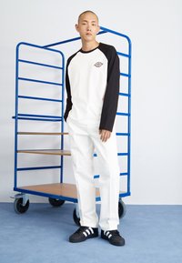 Maglietta a maniche lunghe raglan bianca e nera, abbinata a pantaloni bianchi e scarpe nere con strisce bianche. Sullo sfondo c'è un carrello blu.