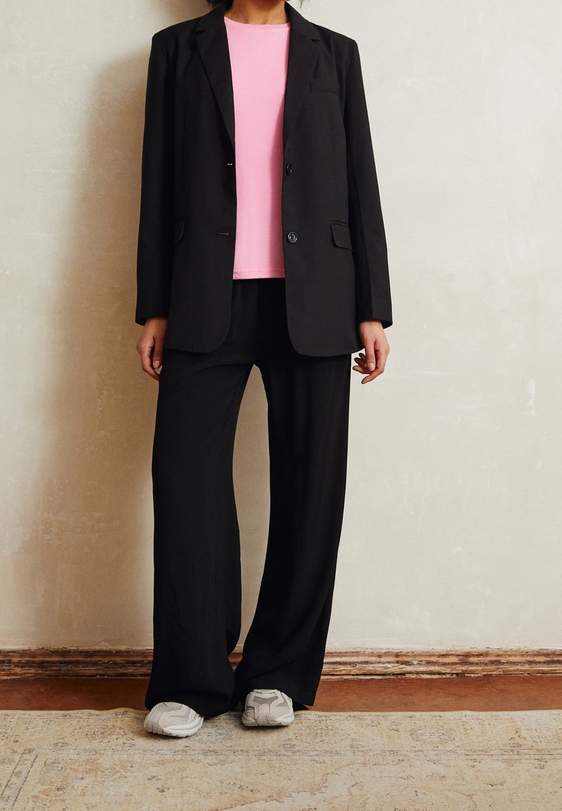 Personne portant un blazer noir et un pantalon à jambes larges avec une chemise rose et des baskets blanches, debout à l’intérieur sur un tapis.