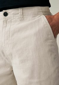 Gros plan sur un pantalon en lin beige avec une main dans la poche avant droite et un bouton noir à la taille.
