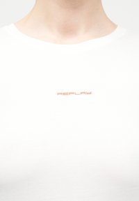 Άνω μέρος στήθους ατόμου που φοράει απλό λευκό μπλουζάκι με μικρό πορτοκαλί λογότυπο "REPLAY" τοποθετημένο στο κέντρο κάτω από το λαιμό.