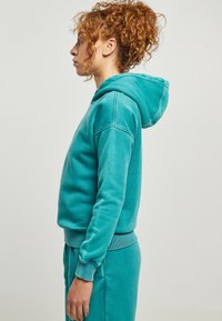 Tirkīza kapuce, kas izgatavota no mīksta auduma, ar brīvu piegriezumu, regulējamu kapuci, ribbelinātām piedurknēm un atbilstošiem apakšējiem apģērbiem.