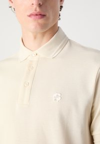 Beige polo shirt med et strukturert mønster, to knapper ved kragen, og en brodert hvit logo på brystet. Glatt stofffinish.