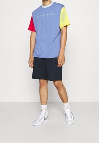 Barevné tričko s krátkým rukávem v modré barvě s červenými, žlutými a zelenými rukávy, s bílým kurzivním logem, doplněné o tmavé šortky a tenisky.