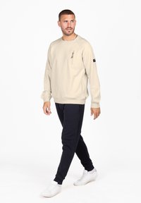 Felpa color beige chiaro con taschino sul petto, abbinata a pantaloni jogger blu scuro e sneakers bianche; vestibilità rilassata e tessuto morbido.