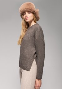 Brązowy sweter dziany z prążkowanym wykończeniem, bocznymi szczelinami i miękką beżową czapką z futra. Długie, falowane włosy okalają twarz, prezentując neutralną paletę kolorów.