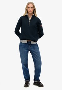 Pull sweater bleu marine à col zippé, avec des poignets et un ourlet côtelés, superposé sur une chemise à rayures bleu et blanc, associé à un jean bleu et des chaussures noires.