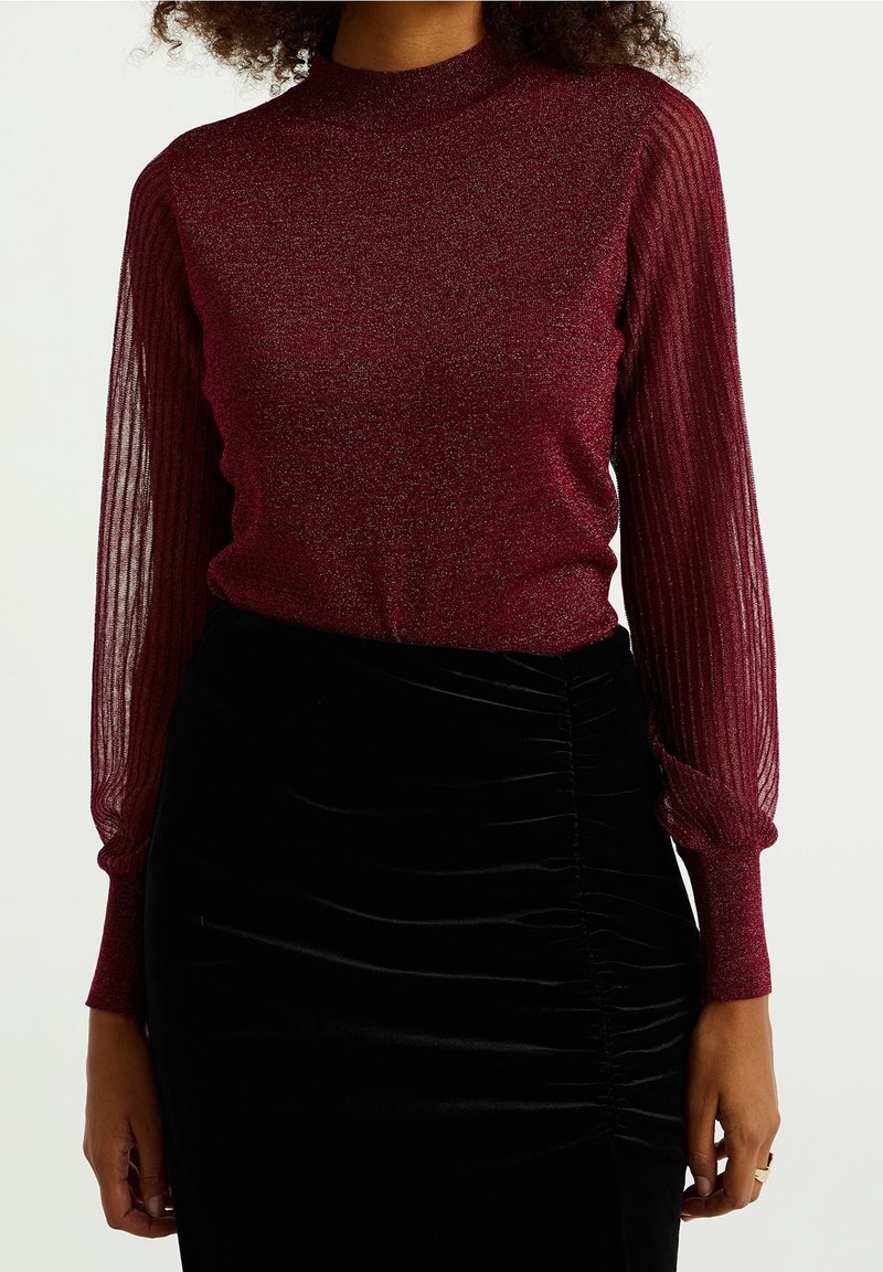 Burgundy långärmad topp med glittrigt huvudtyg och genomskinliga, plisserade ärmar; ihop med en svart, texturerad, draperad kjol.