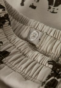 Tejido de algodón beige con cintura elástica y cierre de botón. Presenta una textura arrugada y un estampado gráfico marrón.