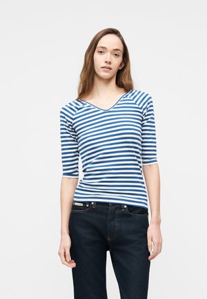 Femme aux longs cheveux bruns portant un haut à rayures horizontales bleues et blanches à manches trois-quarts et un jean foncé, debout devant un fond uni.