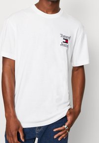 Camiseta de algodón blanca con cuello redondo, que presenta un logo bordado en marino y rojo. Mangas cortas y ajuste relajado. Llevada con jeans azules.