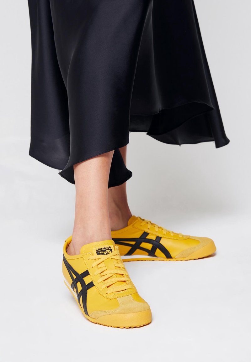 Personne portant une jupe noire et des baskets jaune vif avec des rayures noires debout sur un sol blanc.