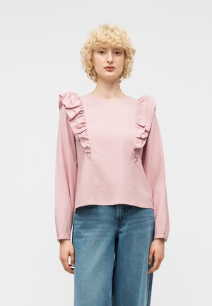 Młoda kobieta z kręconymi blond włosami, ubrana w jasnoróżową bluzkę z długim rękawem i falbanami oraz niebieskie jeansy z wysokim stanem na jednolitym tle.