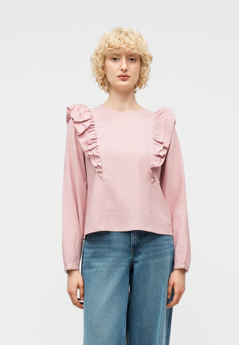 Νεαρή γυναίκα με καστανά σγουρά μαλλιά, που φοράει ανοιχτό ροζ μπλούζα με μακρύ μανίκι και βολάν, καθώς και μπλε τζιν με ψηλή μέση, σε απλό φόντο.