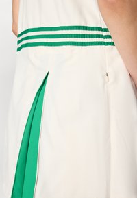 Balta tunika ar zaļiem svītrainiem elastīgiem akcentiem jostasvietā, ar sānu šķirbu, kas atklāj zaļu iekšējo oderi, demonstrējot gludas auduma tekstūras.