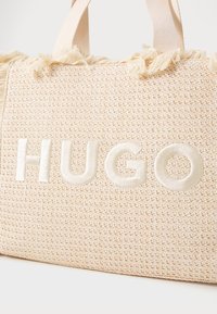 Woven beige tote bag with fringed top, featuring prominent embroidered "HUGO" logo in cream. Textured surface, sturdy fabric handles.