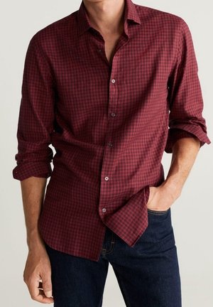 Mężczyzna ubrany w czerwoną i czarną koszulę w kratę z długim rękawem, z podwiniętymi mankietami, oraz ciemnoniebieskie dżinsy, z jedną ręką w kieszeni.