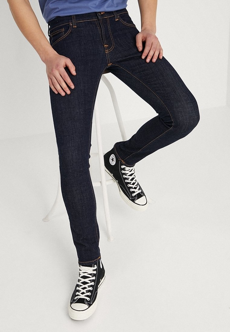 Persona vestita con jeans skinny blu scuro e scarpe alte nere che siede su uno sgabello bianco davanti a uno sfondo semplice.