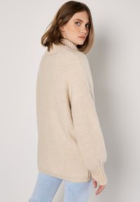 Pull en maille beige, surdimensionné, avec un col montant, des poignets côtelés et un tissu doux et texturé. Il présente des coutures d'épaules tombantes pour plus de confort.