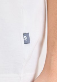 T-shirt en coton blanc avec une petite étiquette grise sur le côté, à manches courtes et à la coupe propre et simple. Texture de tissu doux.