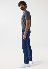 Conjunto masculino que incluye una camiseta de algodón gris y jeans de mezclilla azul. Las zapatillas blancas aportan contraste. Los jeans son de corte ajustado con costuras mínimas.