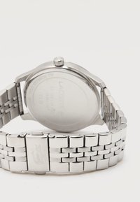 Ασημένιο ρολόι χειρός Lacoste από ανοξείδωτο ατσάλι, εμφανιζόμενο από πίσω με μεταλλικό βραχιόλι και χαραγμένο λογότυπο κροκοδείλου στην πόρπη.