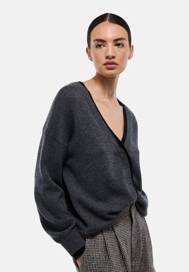 Cardigan en tricot gris avec un décolleté en V profond, manches longues, bord noir et fermeture à boutons, associé à un pantalon à carreaux taille haute.