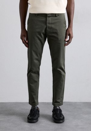 Homme portant un pantalon slim vert foncé et des baskets noires, se tenant devant un fond gris.