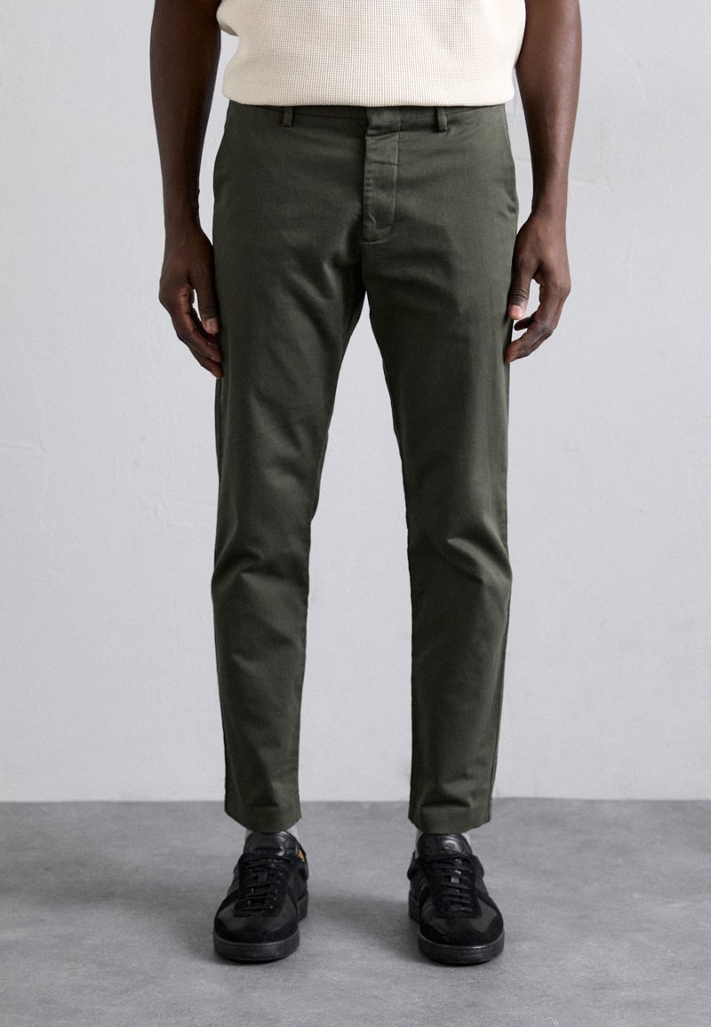 Homme portant un pantalon slim vert foncé et des baskets noires, se tenant devant un fond gris.