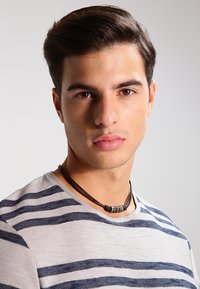 Black leather necklace with round metal accents, worn over a light gray and navy striped shirt, against a plain light background.