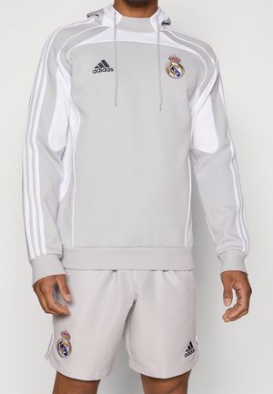 Mężczyzna ubrany w jasno-szarą sportową bluzę z kapturem Adidas Real Madrid oraz pasujące spodenki, z logo marki i klubu na klatce piersiowej i udzie.