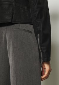 Giacca di pelle nera su pantaloni grigi con una trama liscia, caratterizzati da dettagli con zip sui polsini e una vestibilità elegante e su misura.