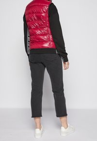 Rode gewatteerde vest met een glanzende afwerking, gedragen over een zwart langemouwen shirt, gecombineerd met grijze cropped jeans en witte sneakers.