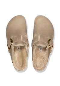 Sabots en daim beige avec une doublure en fourrure douce, une sangle ajustable et un accent de boucle en or. Bout arrondi et semelle intérieure anatomique pour le confort.