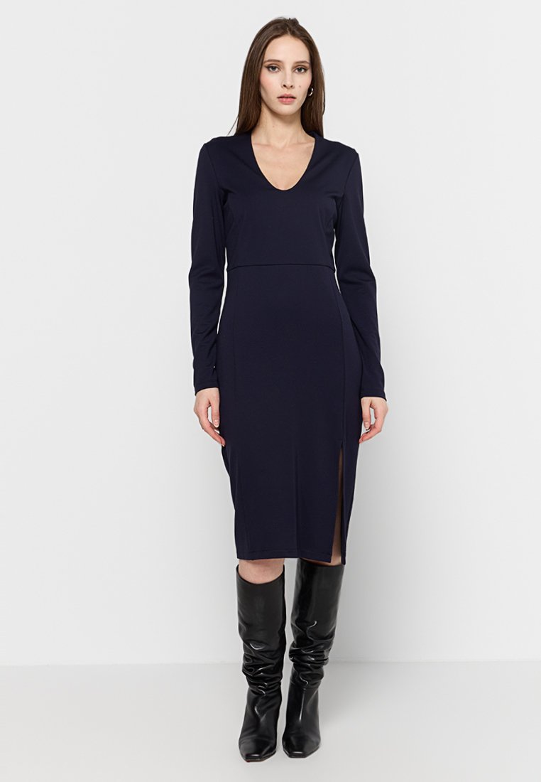 Abito lungo navy a maniche lunghe con scollatura a V, vestibilità aderente, lunghezza fino al polpaccio, spacco laterale, abbinato a stivali neri al ginocchio. Tessuto morbido.
