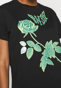 Camiseta negra con estampado gráfico de rosas y hojas en verde y azul claro, con texto estilizado sobre el diseño. Manga corta, cuello redondo.