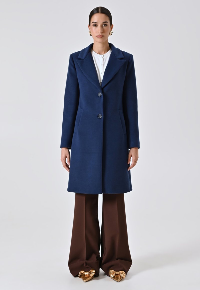 Donna che indossa un cappotto blu navy con bottoni, pantaloni larghi marroni, top bianco e scarpe decorate con fiocchi dorati, in piedi di fronte a uno sfondo neutro.