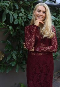 Bordo čipkasta obleka z dolgimi rokavi, okrašena z rastlinskim vzorcem in prilegajočo se silhueto, na ozadju zelene listnate rastline.