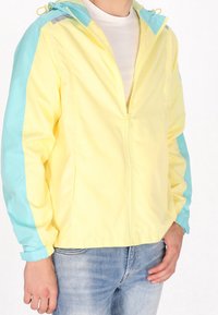 Lichtgewicht windjack met een geel middenstuk en turquoise mouwen, voorzien van een capuchon, ritssluiting en elastische boorden. Gladde nylon textuur.